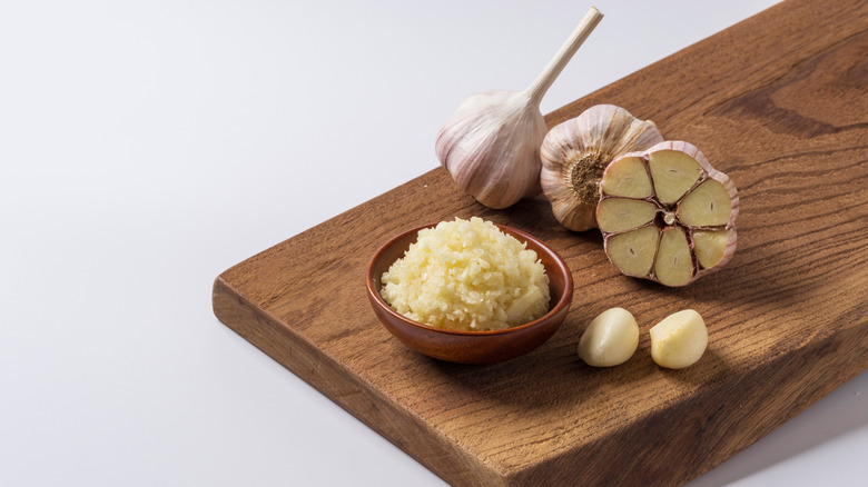 Small bowl of minced garlic on a cutting board, along with cloves and whole garlic