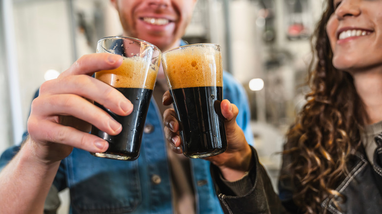 Two people toasting with glasses of dark beer with a blurry brewery background.