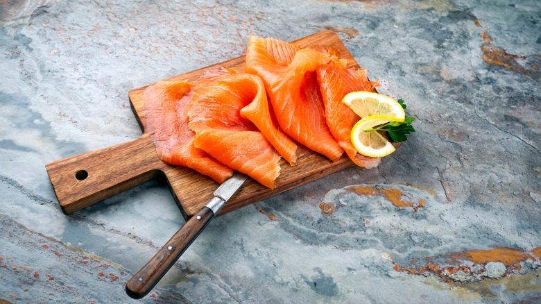 Sliced smoked salmon on a wooden cutting board with a knife and lemon and herb garnish