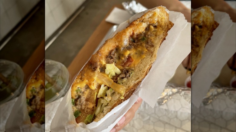 hand holding up chopped cheese sandwich in white wrapper.