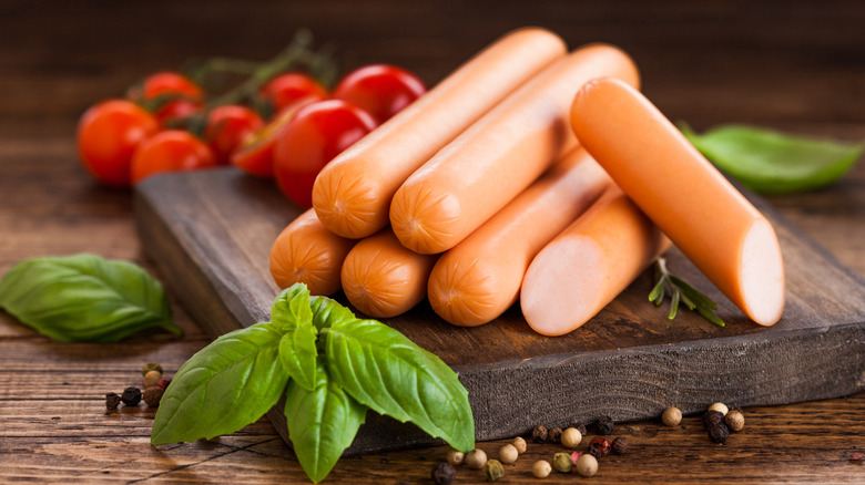 Uncooked hot dogs on wood board with basil and peppercorns in foreground