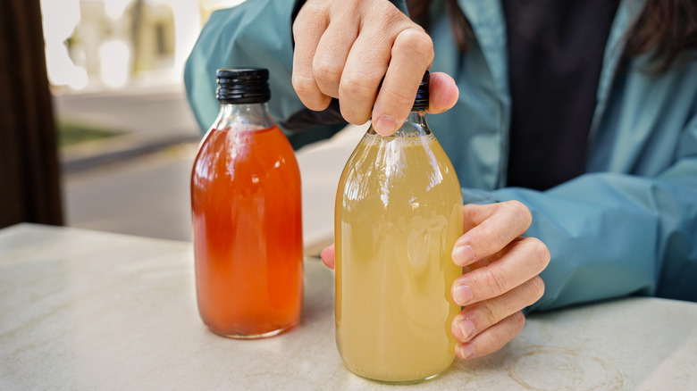 A close-up of two jars of kombucha, one red and one yellow, with someone twisting the cap of the yellow