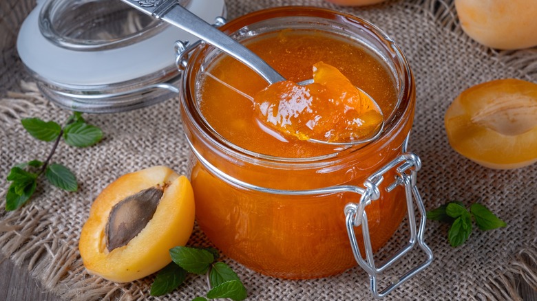 Open jar of apricot jam with halved apricots next to it on woven tablecloth