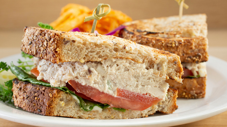A closeup of a tuna salad sandwich with lettuce and tomatoes, with chips in background