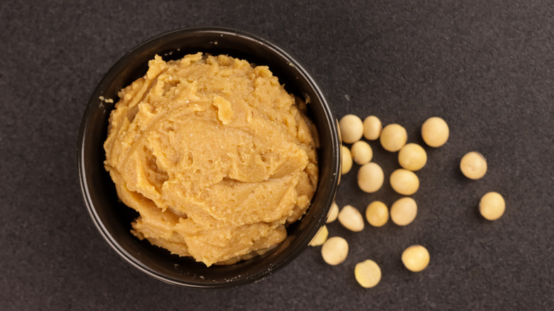 Miso paste in small bowl with soybeans on dark surface, overhead view.