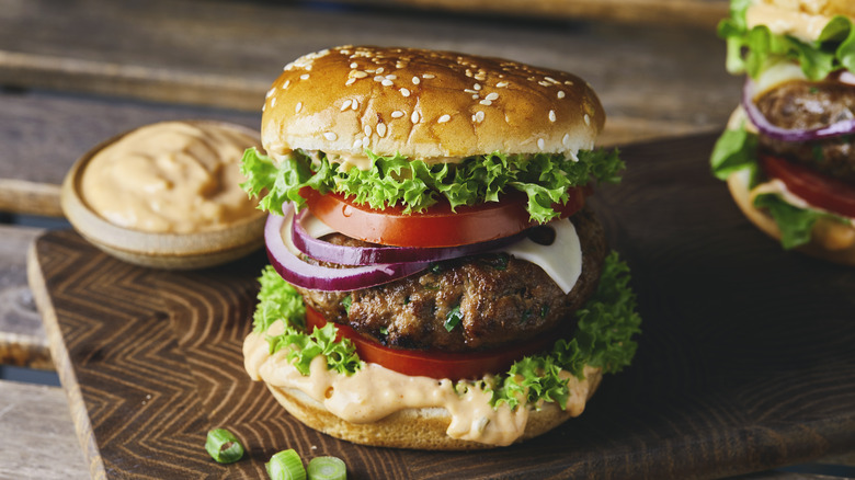 Hamburger with sesame seed bun on wood cutting board surrounded by french fries and tomatoes