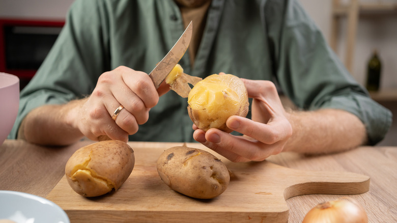Someone peeling potatoes with a knife over a wooden cutting board