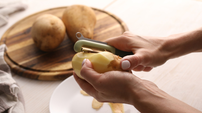 Someone using a vegetable peeler to peel a potato, with two other potatoes in the background