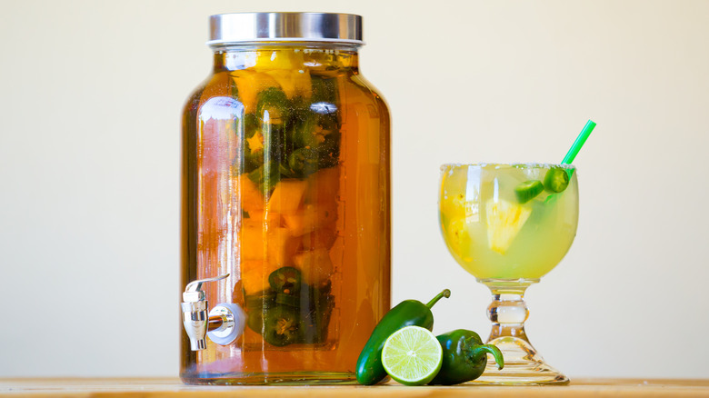 Tequila infused with jalapeños in a large jar with whole jalapeños in front and a margarita on the side