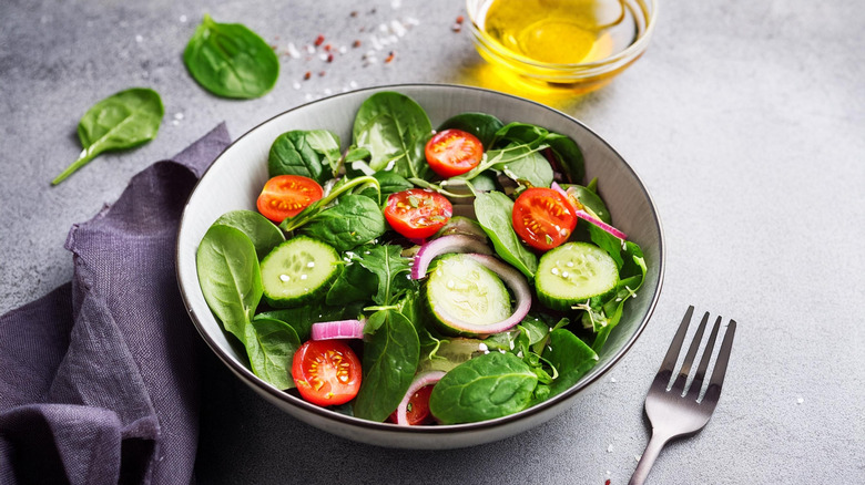 A bowl of salad with spinach, tomatoes, cucumber, and red onion, with salad dressing in a glass bowl in the background