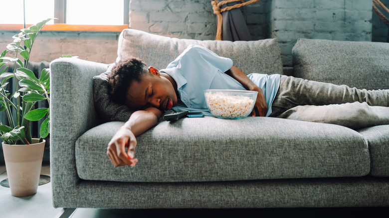 A man asleep on a gray couch with a bowl of popcorn and a TV remote