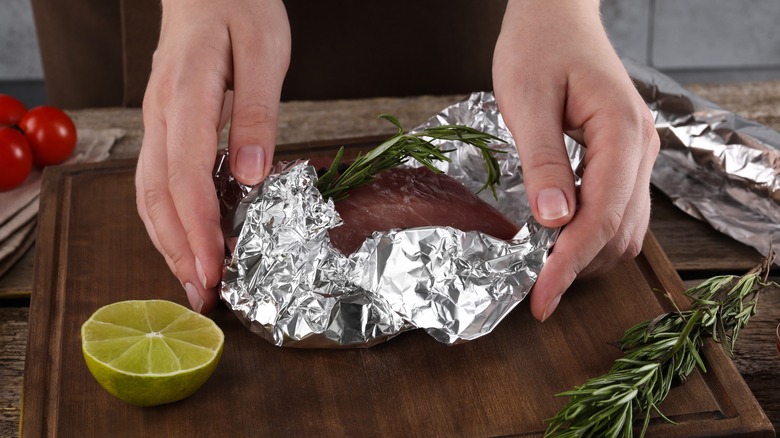 Close-up of someone wrapping meat with herbs in aluminum foil