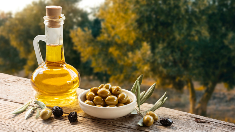 A close-up of a small white bowl of olive sitting on a wooden railing, with a corked glass bottle of olive oil to the left, both overlooking a field of olive trees