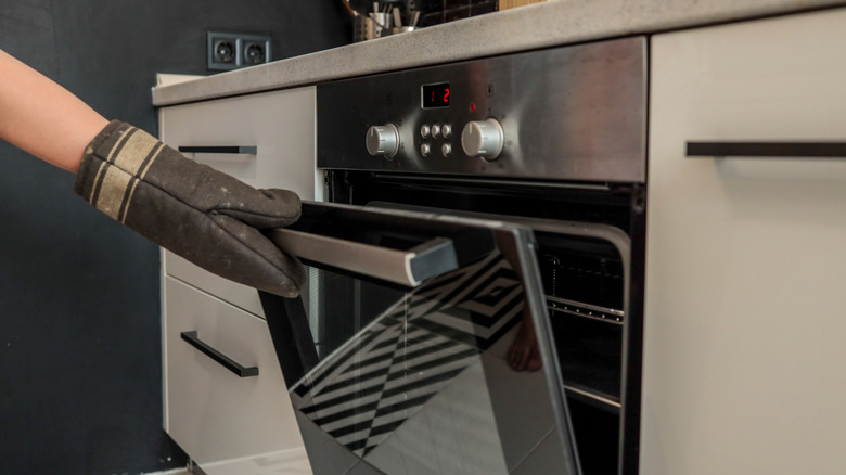A close-up of someone opening an oven door while holding a dark brown oven mitt