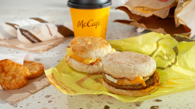 McDonald's breakfast foods including hash browns, coffee, an egg mcuffin and a breakfast biscuit sandwich laid out on a table with an open paper bag in the background