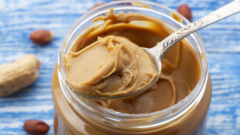 Close-up of a spoonful of peanut butter on an open jar, with peanuts on a blue table