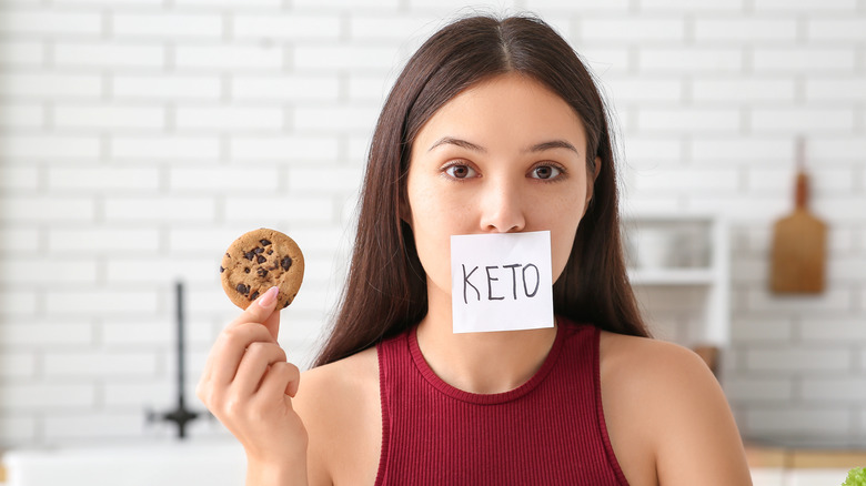 a young woman with a piece of paper over her mouth reading "Keto" holding a cookie