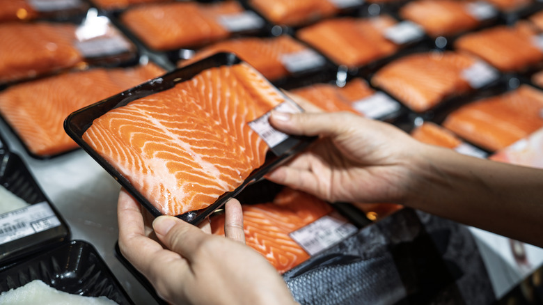 Closeup of hands picking up packaged salmon filet from cold case.