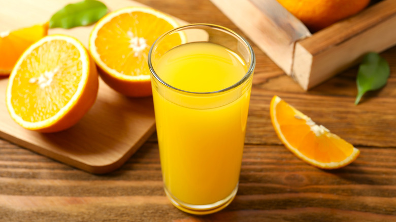Glass of orange juice on wood surface, cut oranges in background.