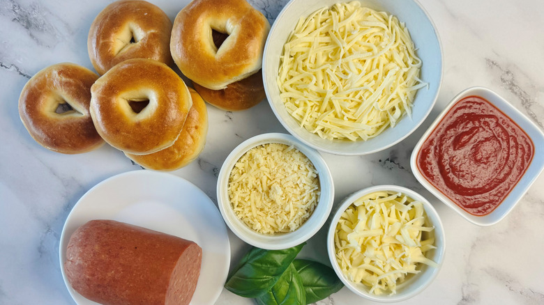 mini bagels, pepperoni, shredded cheeses in bowls, pizza sauce, and basil leaves laid out on marble surface