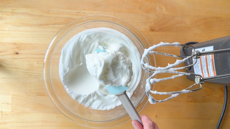 Stiffly beaten egg whites in glass bowl with hand mixer and rubber spatula