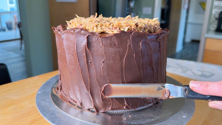 Using an offset spatula to spread chocolate ganache on the sides of a German chocolate cake