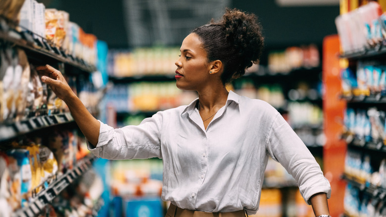 Woman in grocery aisle reaches for product on a shelf.