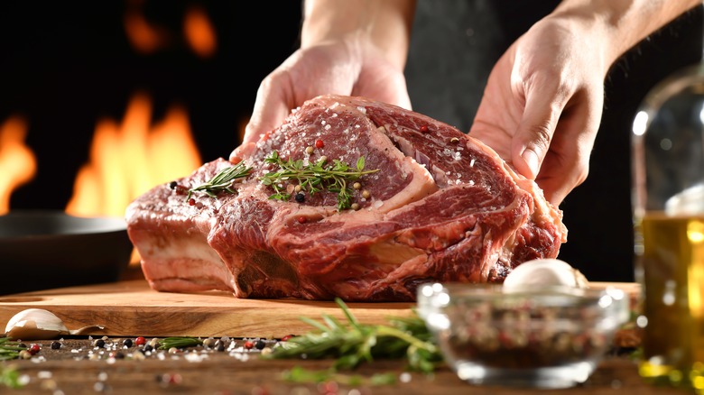 Chef preparing raw ribeye steak on cutting board with seasonings.