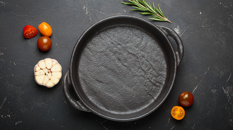 Overhead shot of cast iron pan surrounded by garlic, rosemary and tomatoes on dark background.