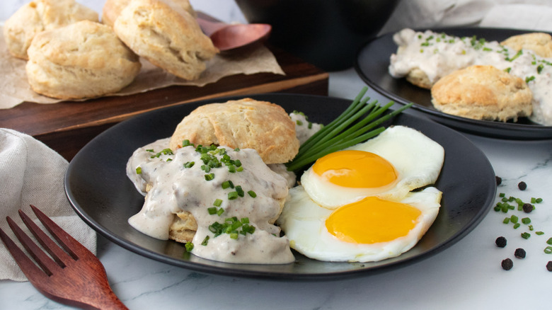 Side shot of a plate of biscuits and gravy with chives and fried eggs on the side