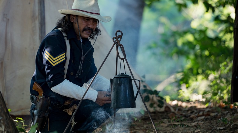 man in a cowboy hat brews coffee over smoldering fire