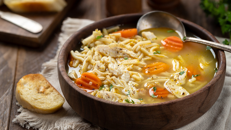 A bowl of homemade chicken noodle soup with a spoon and a small slice of toasted bread next to it