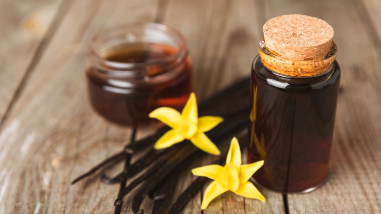 A bottle and bowl of vanilla extract next to vanilla bean pods and flowers
