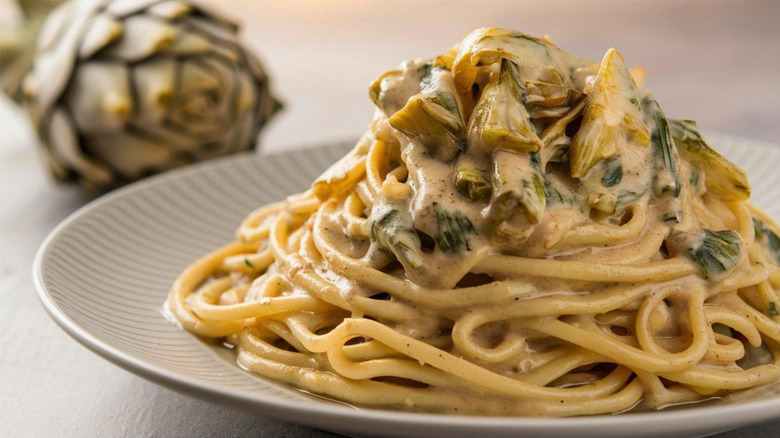 A plate of pasta with artichoke in a cone shape on a white plate, with a whole artichoke in the background
