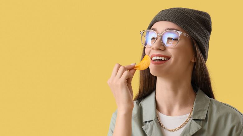young woman wearing a beanie and glasses smiling and eating a potato chip on a yellow background