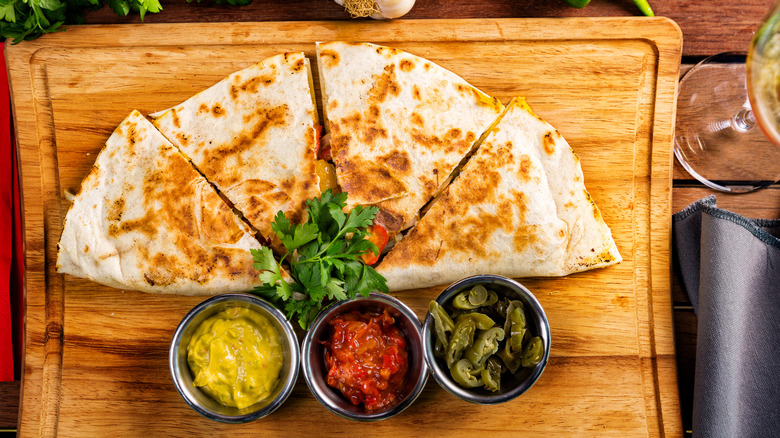 An overhead shot of a quesadilla split into four pieces on a cutting board, with toppings in small metal bowls next to it