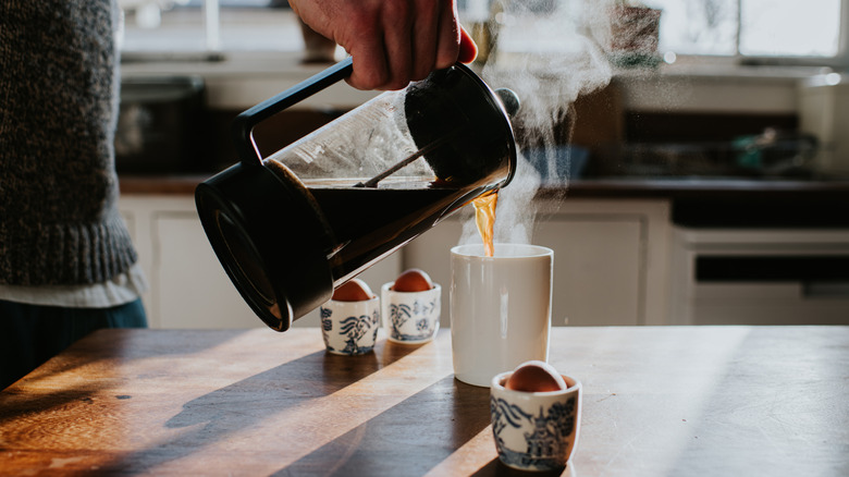 A close-up of someone pouring coffee from a French press into a white coffee mug, with egg cups sitting around the mug