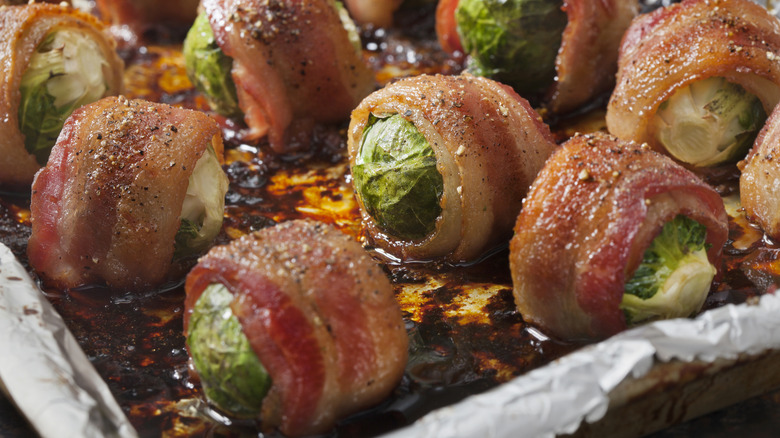 A close-up of bacon-wrapped Brussel sprouts cooking on a pan covered in aluminum foil