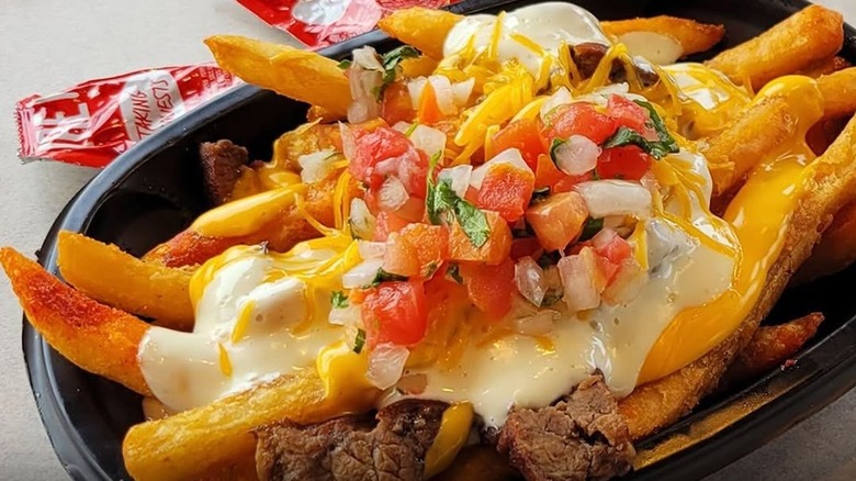 A close-up of a black plastic tray filled with Taco Bell's steak garlic nacho fries, with two used sachets of sauce behind it