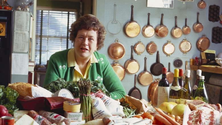 Julia Child in kitchen with ingredients and copper pans on wall