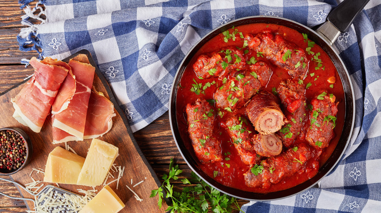 rolled beef braciole in a pan with tomato sauce and herbs, prosciutto and parmesan cheese on the side.