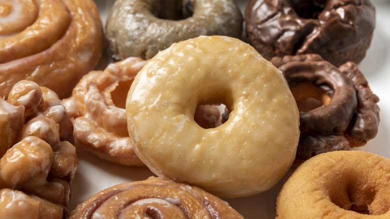 Overhead photograph of an assortment of donuts on a white background. Glazed, cake, and chocolate covered donuts as well as a cinnamon roll and specialty donuts are arranged in a box that is sitting on a white background.