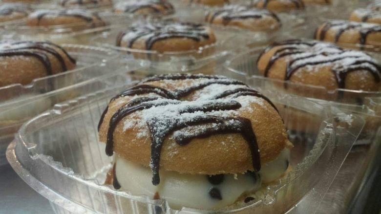 Multiple plastic containers of cannoli donuts sliced in half, filled with cream, and drizzled with chocolate