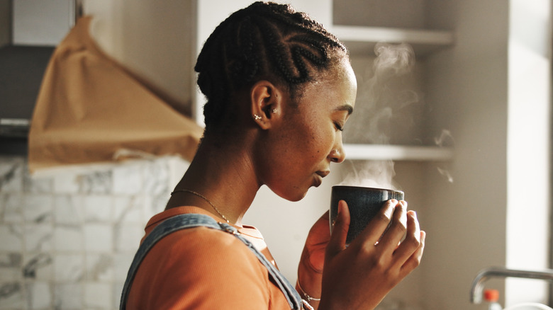 Woman stands in kitchen while smelling cup of coffee