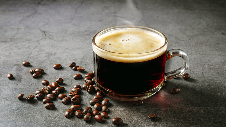 A close-up of a steaming glass mug of coffee, with coffee beans scattered around it