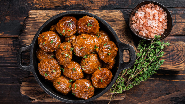 Cooked meatballs in a cast iron skillet on a wooden cutting board, with a black ramekin of pink Himalayan sea salt and fresh herbs