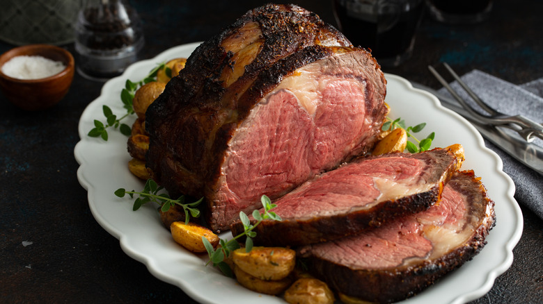 A platter of cooked prime rib with two slices on a white serving tray, surrounded by potatoes