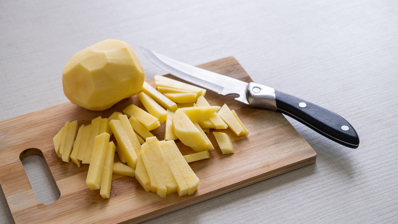 A cut-up potato sits on a wooden board with a knife
