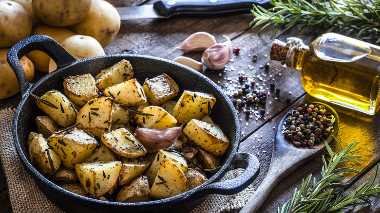 Fancy cooked diced potatoes in a cast iron skillet, with whole potatoes, a knife, and some herbs in the background, and a bottle of olive oil and a spoonful of peppercorn to the side