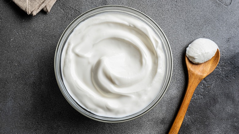 Overhead view of Greek yogurt in glass bowl with wooden spoon, dark background.